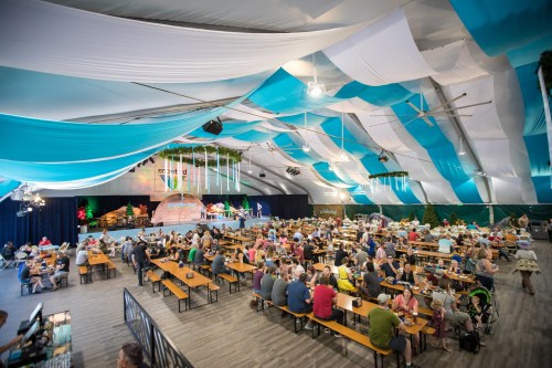 A large indoor event space filled with people dining at long tables under a colorful, draped ceiling.