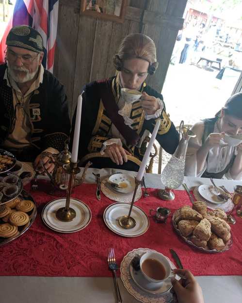 A historical tea gathering with three people, a table set with pastries, tea, and candles in a rustic setting.