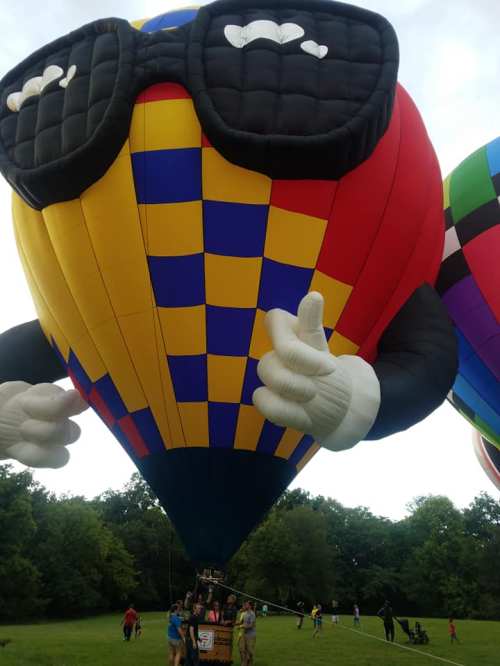 A colorful hot air balloon with sunglasses and cartoonish hands stands in a grassy field, surrounded by people.
