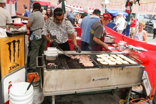 A chef grills meat and toasts bread at a busy outdoor food festival, surrounded by people and colorful tents.