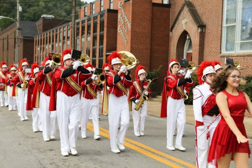 A marching band in red and white uniforms parades down the street, with musicians playing instruments and a dancer in red.