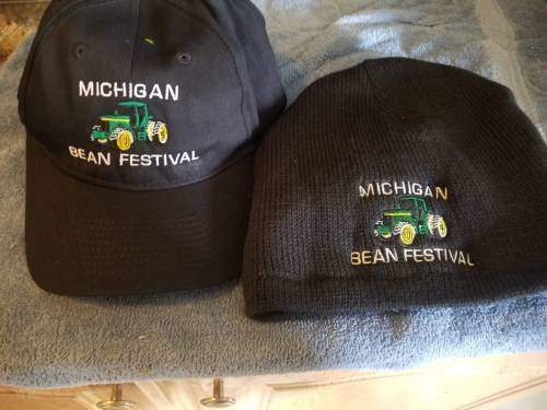 A black baseball cap and a black beanie, both featuring "Michigan Bean Festival" and a green tractor design.