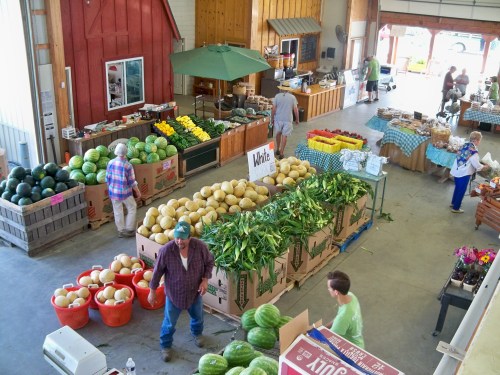 A bustling farmers market with various fruits and vegetables displayed in crates and baskets, and shoppers browsing.