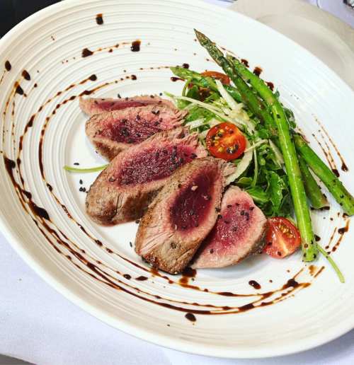 A plate of sliced steak with a pink center, served with asparagus, cherry tomatoes, and a drizzle of balsamic reduction.