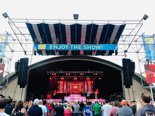 A crowd watches the Newsboys perform on stage under a striped canopy, with a banner reading "Enjoy the Show!" above.
