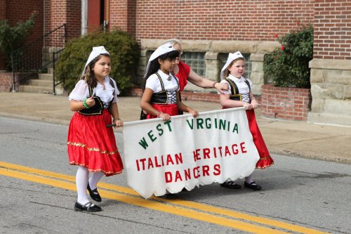 Three young girls in traditional costumes hold a banner reading "West Virginia Italian Heritage Dancers" during a parade.