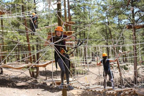 A group of people in helmets navigating a ropes course in a forested area, smiling and climbing on ropes and platforms.