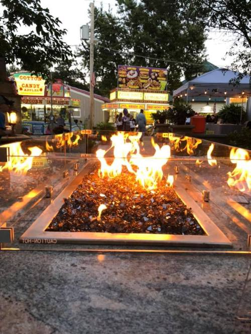 A fire feature with flames rising from glass stones, set against a backdrop of a fair with colorful food stalls.