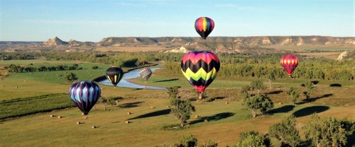 Colorful hot air balloons float over a scenic landscape with rolling hills and a winding river.