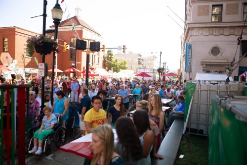 A large crowd gathers at a street festival, with people enjoying food and activities under sunny skies.