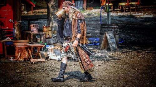 A man in medieval attire walks through a rustic setting, sipping from a mug, with various props in the background.
