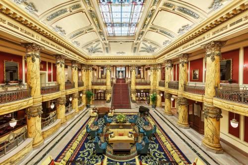 Luxurious grand hall with marble columns, ornate ceiling, and elegant furnishings, featuring a large staircase and colorful rug.