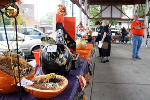 A festive display of decorated pumpkins and autumn decorations at a community event, with people browsing nearby.