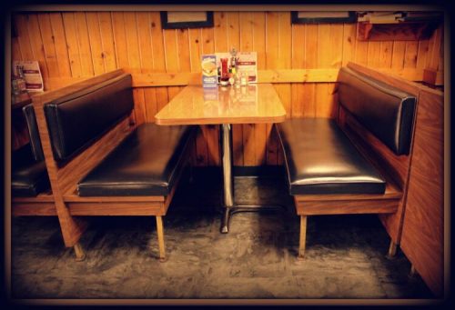 A cozy diner booth with a wooden table and black leather seating against a wooden wall.