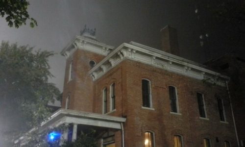 A historic brick building shrouded in fog, with a tower and decorative roof details, illuminated by blue light.