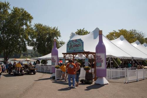 A lively outdoor wine pavilion with large wine bottle decorations and a tent, bustling with visitors enjoying the festival.