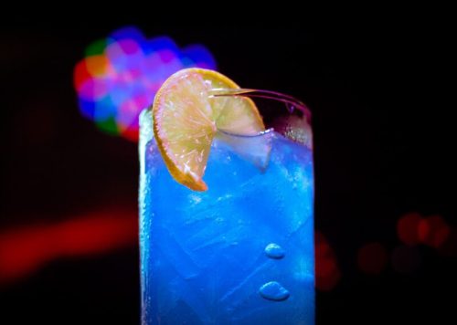 A vibrant blue cocktail with ice and a slice of lemon, set against a colorful, blurred background.