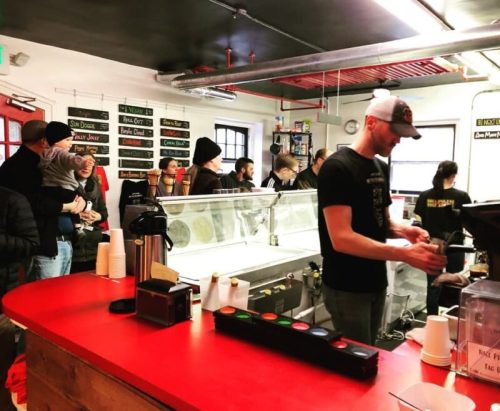 A busy café with a red counter, a barista serving customers, and people waiting in line. Ice cream display in the background.