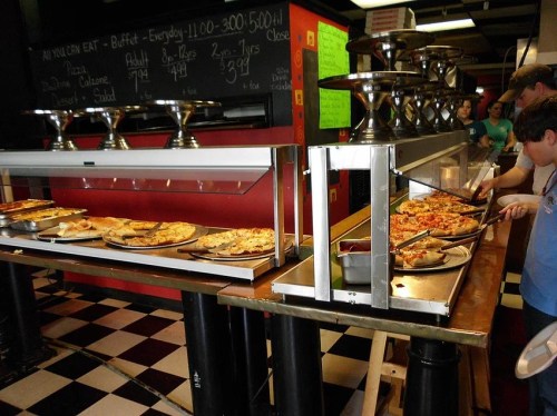 A buffet with various pizzas and pasta dishes, with people serving themselves in a casual dining setting.