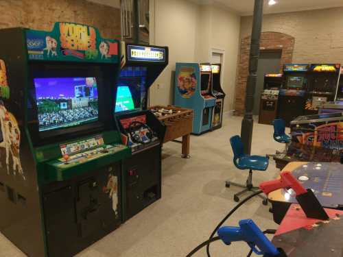 A retro arcade room featuring various classic arcade machines and a pinball table, with blue chairs scattered around.
