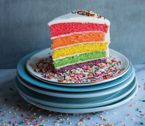 A slice of colorful layered cake with pink, orange, yellow, green, and purple layers, topped with white frosting and sprinkles.