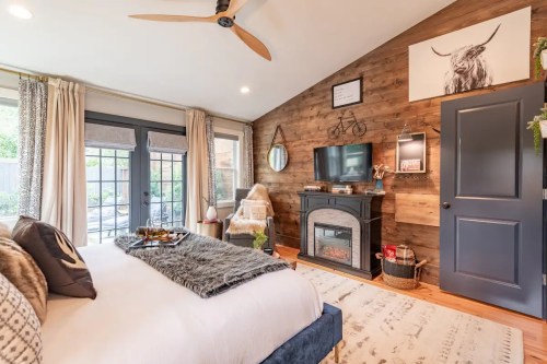 Cozy bedroom with wooden accents, a fireplace, and large windows overlooking a patio. Decor includes a wall-mounted TV.