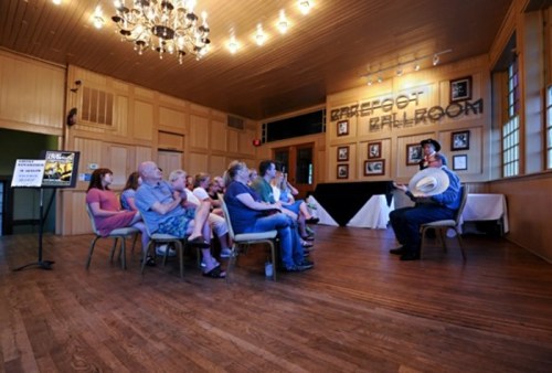 A speaker in a hat addresses an audience seated in a wooden ballroom with framed photos on the walls.