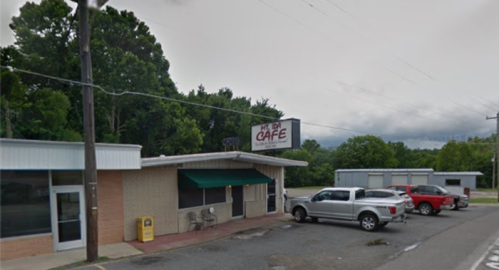 A small café building with a sign, surrounded by trees and parked cars on a cloudy day.