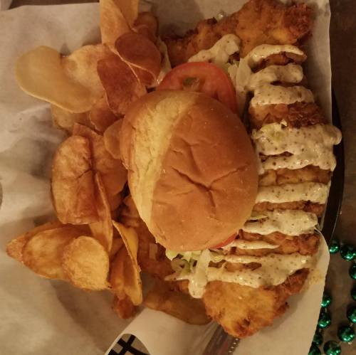 A plate with a fried chicken sandwich, drizzled with sauce, and a side of crispy potato chips.