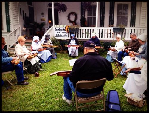 A group of people playing dulcimers outdoors, seated in a circle on a grassy area near a house.