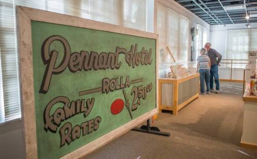 A vintage sign for the Pennant Hotel with family rates, displayed in a museum setting with two people in conversation.