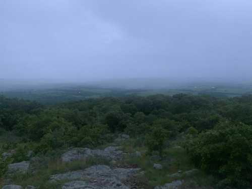 A misty landscape view from a hilltop, with rolling green hills and trees under a cloudy sky.