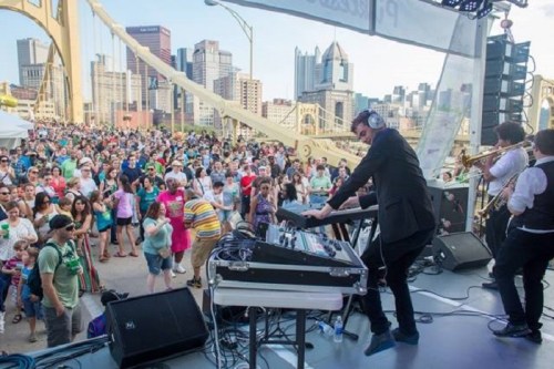 A lively outdoor concert on a bridge, with a crowd enjoying music and performers on stage in Pittsburgh.