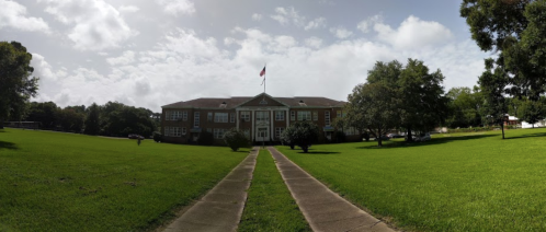 A large brick school building with a flag, surrounded by green grass and trees under a partly cloudy sky.