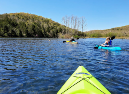 Two people kayaking on a calm lake surrounded by green hills and trees under a clear blue sky.