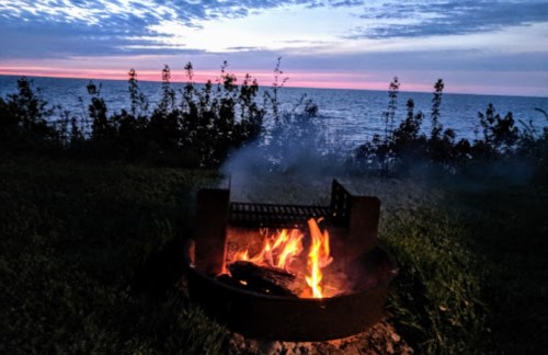 A campfire burns in a grill by the lake at sunset, with trees silhouetted against the colorful sky.