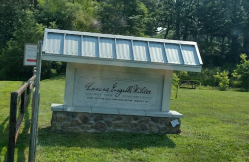 Sign for Laura Ingalls Wilder, featuring a white structure with a metal roof, surrounded by greenery.
