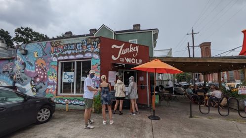 A colorful restaurant exterior with a mural, featuring people waiting in line and outdoor seating under orange umbrellas.