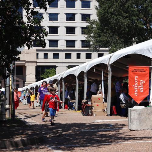 A bustling outdoor book festival with tents, people walking, and a sign for "Authors Alley" in a sunny setting.