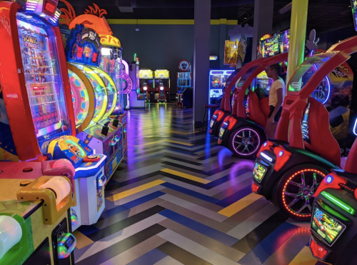 A vibrant arcade filled with colorful games and neon lights, featuring racing machines and various arcade cabinets.