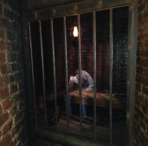 A dimly lit jail cell with brick walls, featuring a barred door and a figure sitting on a wooden bench inside.
