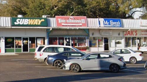 A row of storefronts in a shopping plaza, featuring Subway, The Sicilian II, Mr. Greek, and Jak's, with parked cars in front.