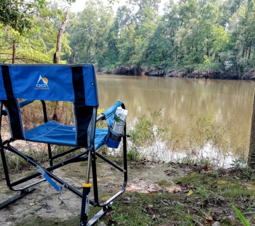 A blue camping chair sits by a calm river, surrounded by trees and greenery on a sunny day.