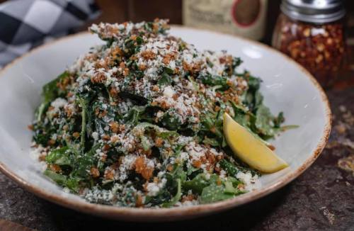 A bowl of green salad topped with breadcrumbs, cheese, and a lemon wedge, with a jar of red pepper flakes in the background.