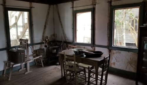 Interior of a rustic room with a wooden table, chairs, and windows, featuring peeling walls and simple furnishings.