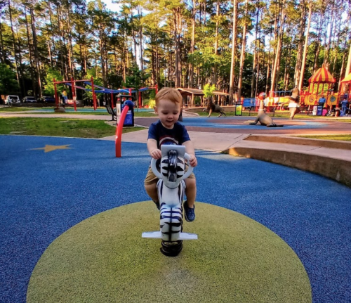A young child joyfully rides a toy motorcycle on a colorful playground surrounded by trees and play structures.