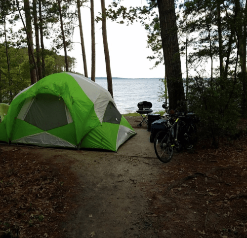 A green and gray tent set up near a lake, with a grill and bicycle nearby, surrounded by trees.