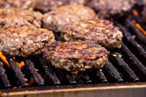 Grilled beef patties sizzling on a barbecue grill, with flames visible beneath.