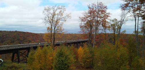 A scenic view of a wooden bridge surrounded by vibrant autumn foliage and rolling hills under a cloudy sky.