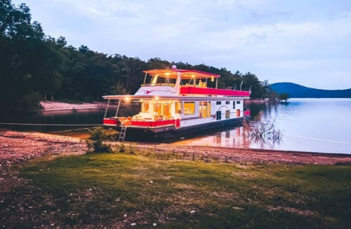 A colorful houseboat docked by a serene lake at dusk, surrounded by trees and hills in the background.